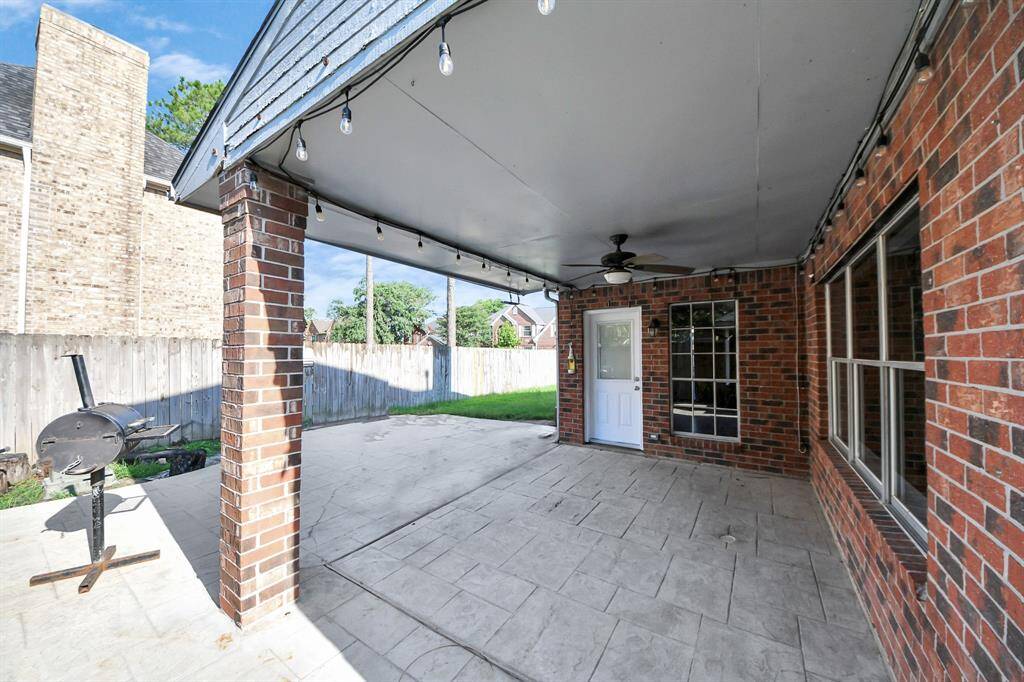 This photo showcases a covered patio with a brick exterior, featuring a ceiling fan and string lights. The space opens to a small yard with a wooden fence, and includes a barbecue grill, ideal for outdoor entertaining.