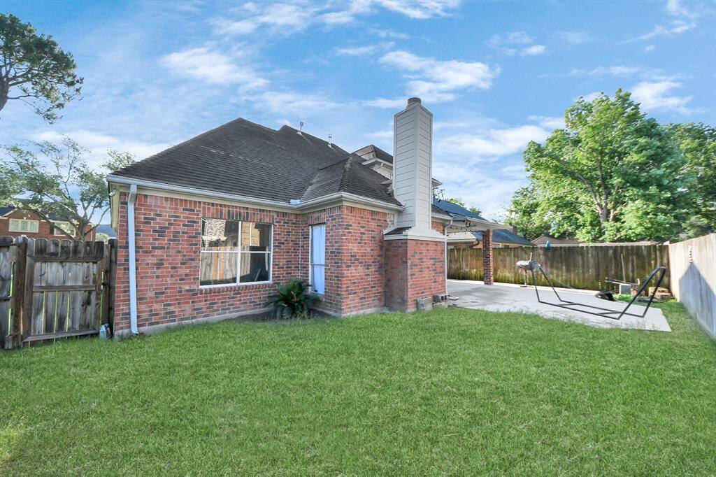 This photo showcases a charming brick home with a spacious backyard. The yard features a well-maintained lawn, a concrete patio area with a swing set, and a wooden fence for privacy. The exterior includes a chimney and a side window, offering a cozy and inviting atmosphere.