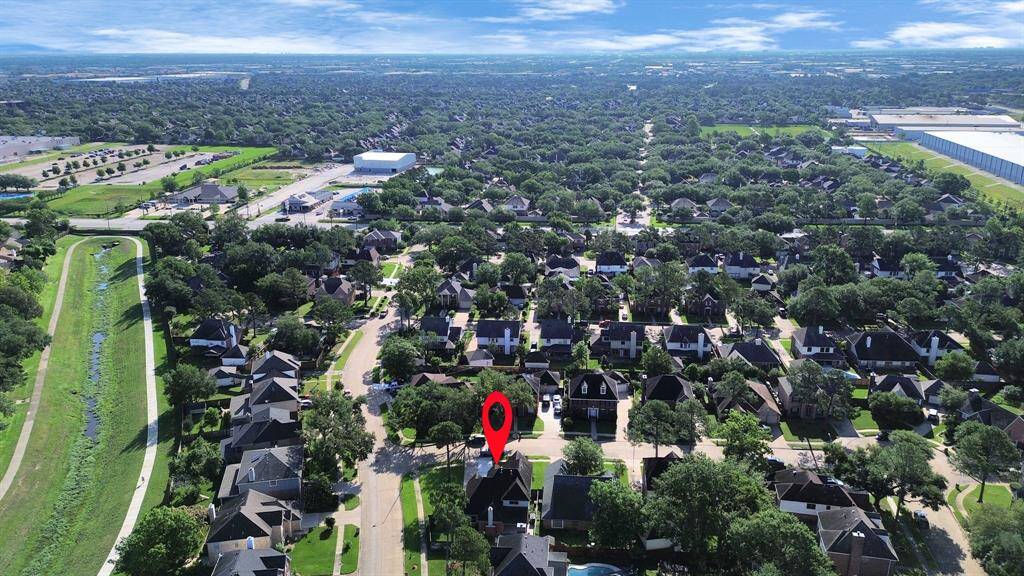 This aerial photo showcases a suburban neighborhood with tree-lined streets and well-maintained homes. The area features a nearby walking path alongside a grassy canal, offering scenic outdoor space. The location is marked by a red pin, highlighting its position within this peaceful community.