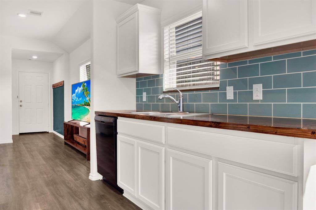 This modern kitchen features white cabinetry, a wooden countertop, and a stylish blue subway tile backsplash. Natural light filters through a window above the sink, and there’s an adjacent space leading to an entrance, providing a welcoming and functional setup.