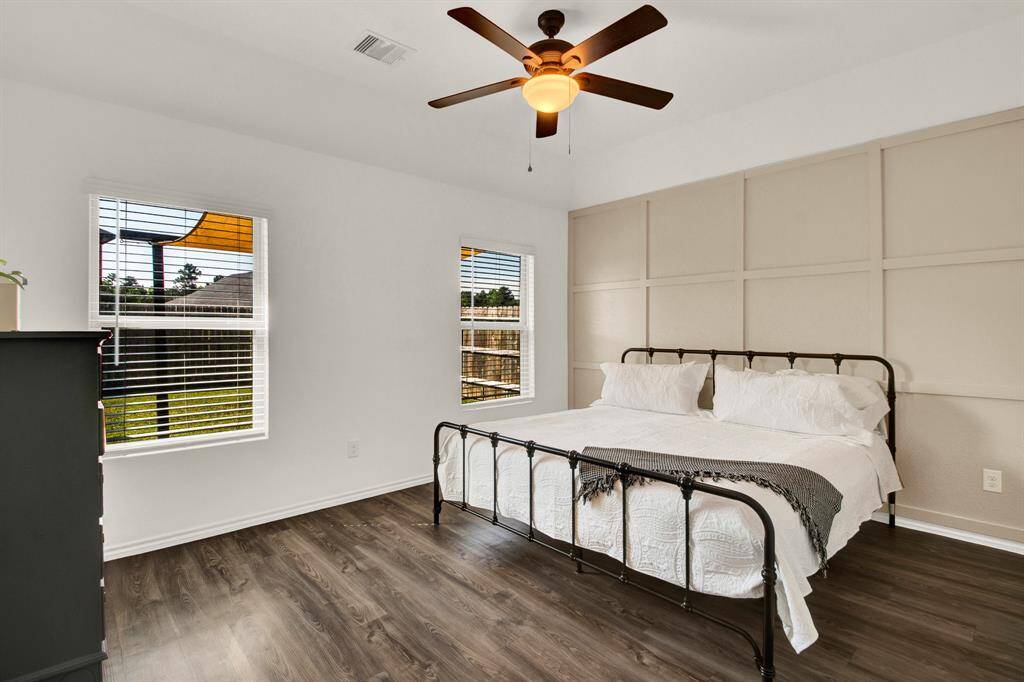 This bedroom features a clean, modern design with a neutral color palette. It has large windows for natural light, a ceiling fan and hardwood-style vinyl flooring.