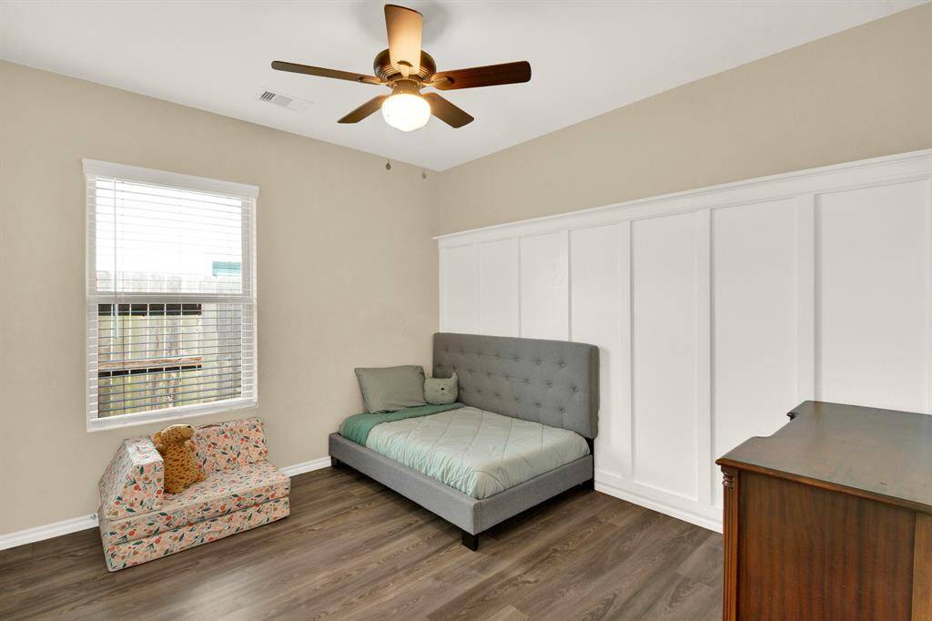 This bedroom features a cozy setup with elegant wainscoting on one wall, a ceiling fan, and a large window providing natural light. The room has wood-look vinyl flooring creating a warm and inviting space.