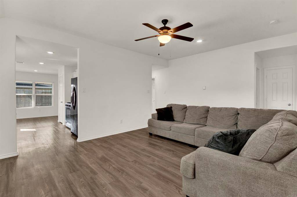 Spacious living area with hardwood-style vinyl flooring and a ceiling fan. The open layout leads to the kitchen, which features black appliances and ample natural light from large windows.