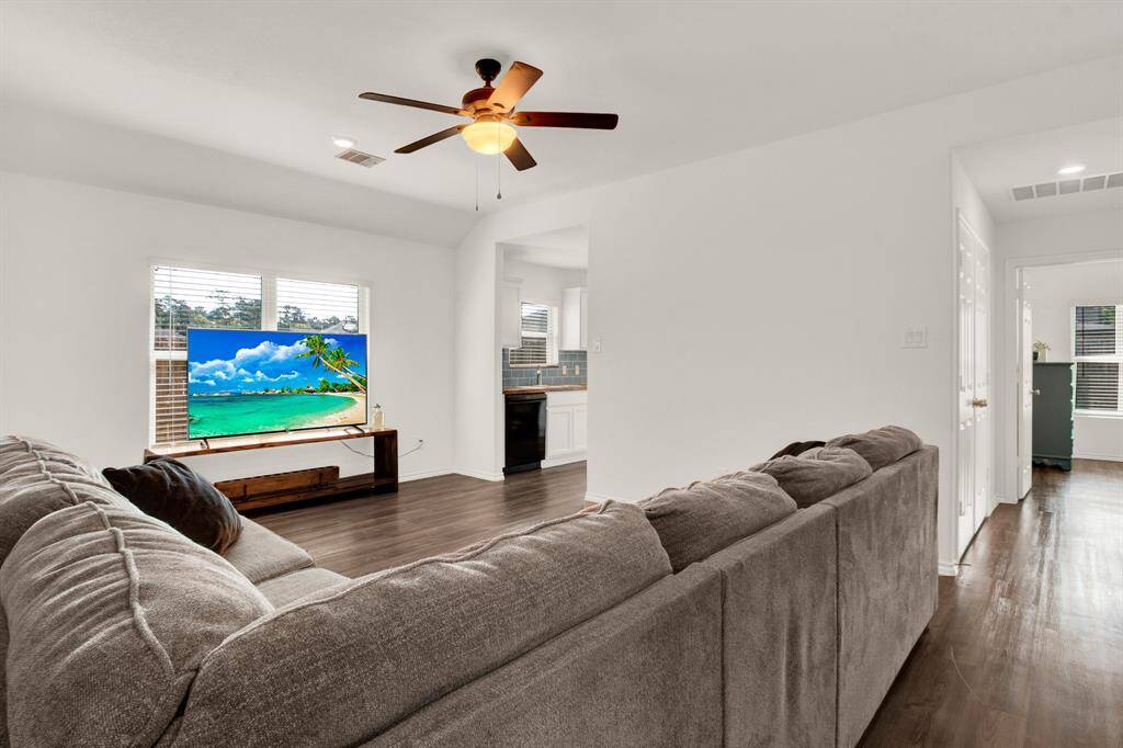 Spacious living area with hardwood-style vinyl flooring, neutral walls, and a ceiling fan. Features an open layout leading to the kitchen, and ample natural light from large windows.