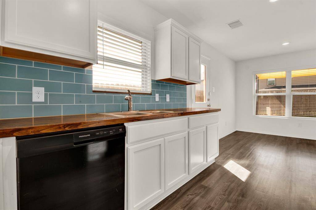 Bright kitchen with modern white cabinetry, wood countertops, and a stylish blue subway tile backsplash. It features a built-in dishwasher and large windows for natural light, complemented by hardwood-style vinyl flooring.