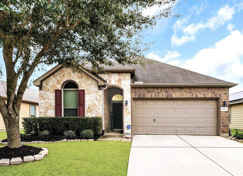 This charming single-story home features a clean, modern facade with brick and stone accents, a neatly manicured front yard, and a spacious two-car garage. The large tree provides ample shade, enhancing its curb appeal.