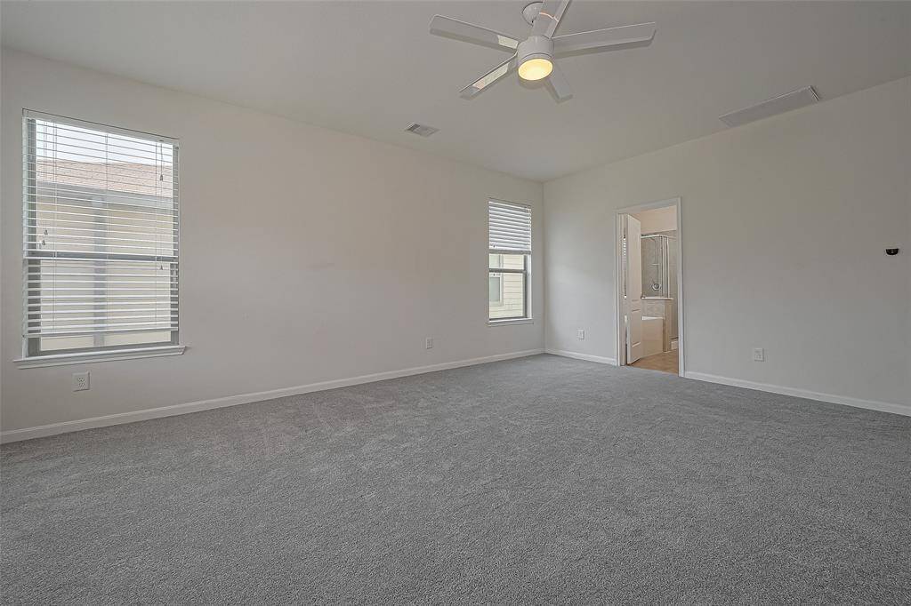 This spacious bedroom features soft carpeting, neutral walls, and a ceiling fan. It has ample natural light from two windows and an en-suite bathroom for added convenience.
