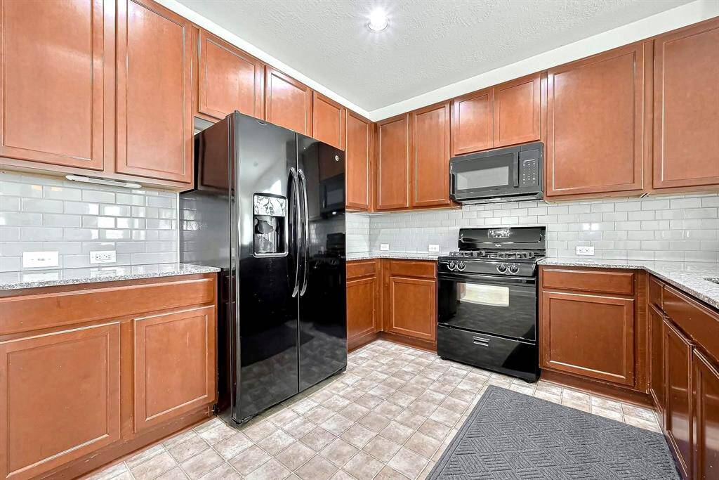 This kitchen features black appliances, including a refrigerator and stove, set against rich wooden cabinetry. The countertops are granite. The space is well-lit, enhancing its modern and functional appeal.
