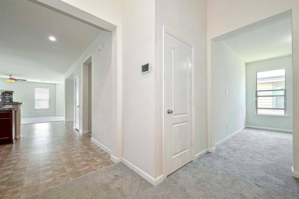 This photo shows a bright and inviting hallway with light-colored walls and a mix of tile and carpet flooring. It leads to an open living area with large windows and a kitchen featuring dark cabinetry. The space is well-lit and modern.