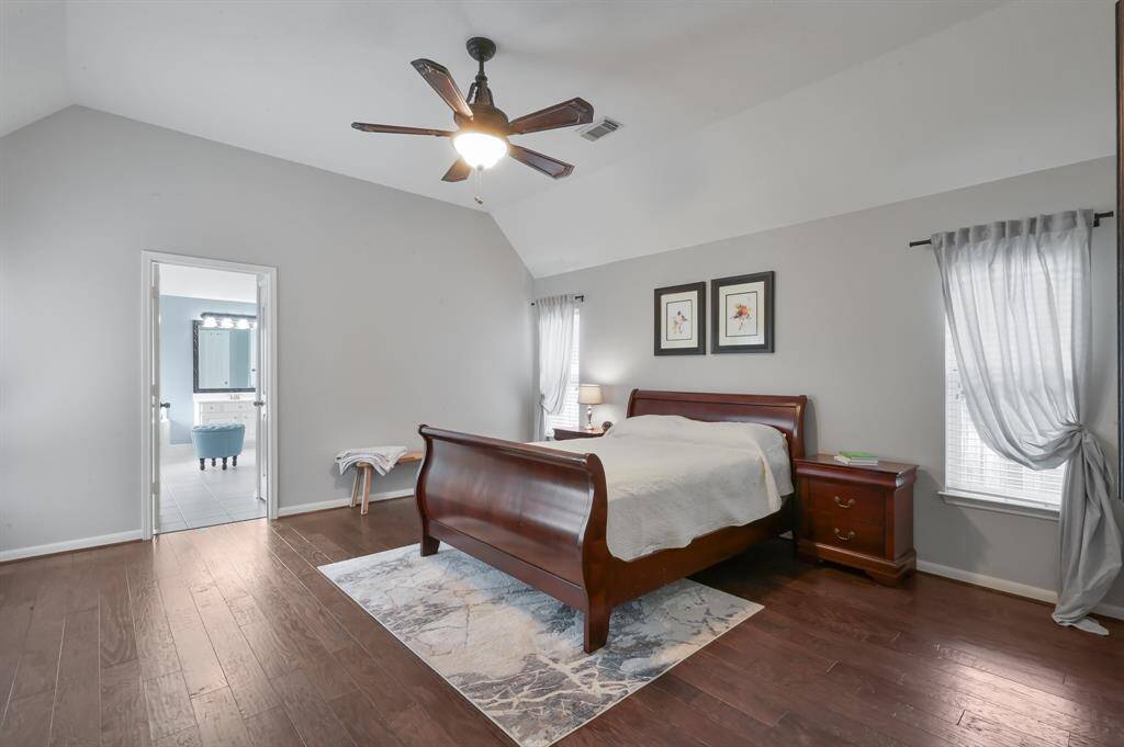 The primary bedroom features a vaulted ceiling with a modern ceiling fan, hardwood floors and ample natural light from large windows and sheer curtains.