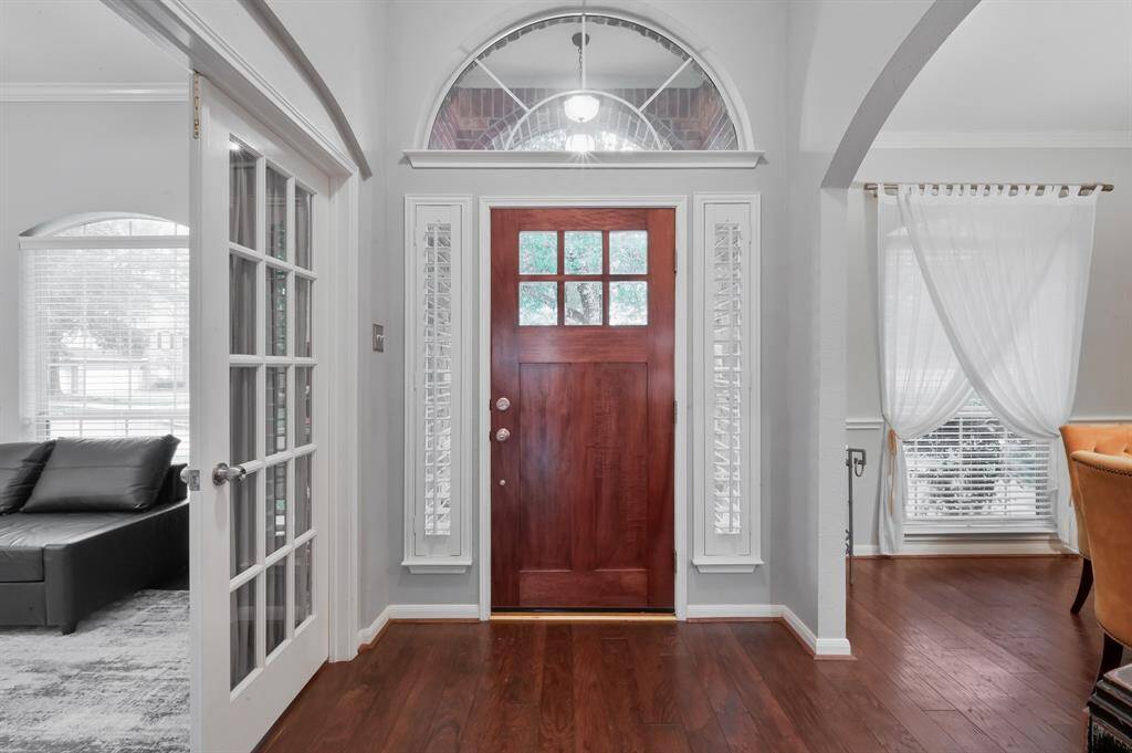 A classic solid wood door with decorative glass panels and sidelights with wooden shutters provide a welcoming entry adjacent to the office/flex room and dining area on right, accented by elegant drapes.