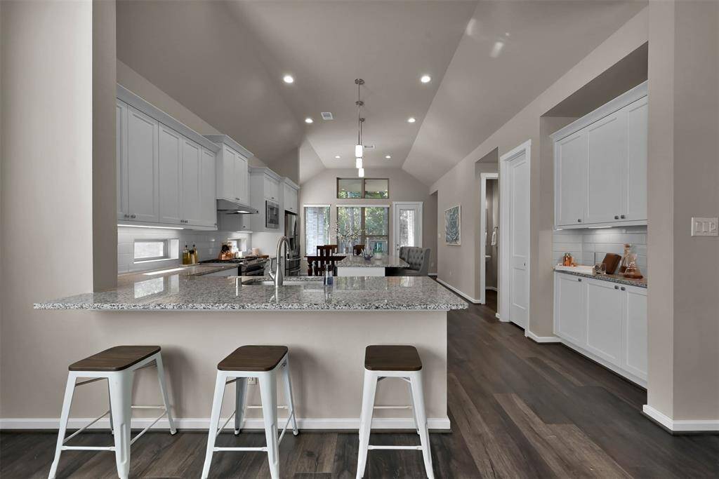 This photo showcases a modern, open-concept kitchen and living area. It features a spacious island with granite countertops and bar seating, sleek white cabinetry, stainless steel appliances, and rich wood flooring. The area is well-lit with recessed lighting and has a view of a dining space and seating area in the background.