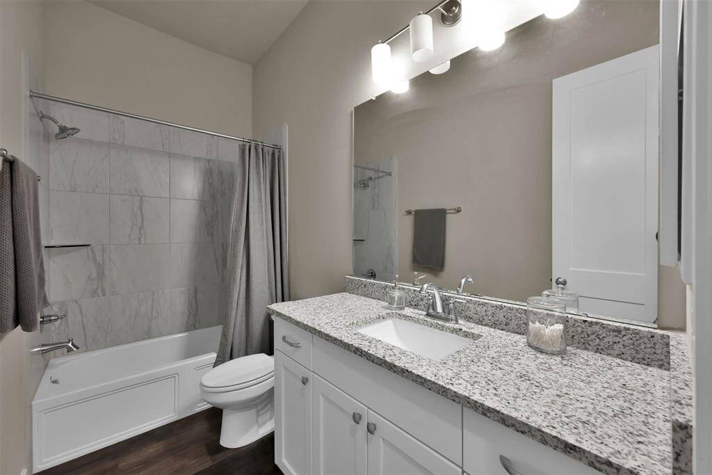 This secondary bathroom features a modern design with a granite countertop, a single under-mount sink, and ample cabinet storage. It includes a tiled shower-tub combo, a large mirror, and elegant lighting, offering both functionality and style.