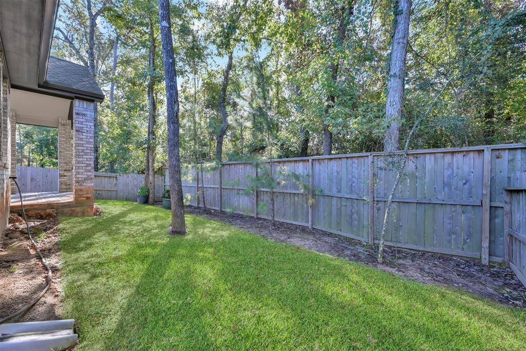 This backyard features a lush, green lawn bordered by a wooden fence, offering privacy and natural views with surrounding trees. A side view of the house shows a brick exterior and a covered patio area, perfect for outdoor relaxation.