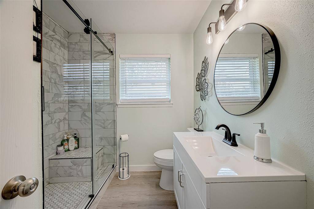 This bathroom features a modern design with a sleek vanity, round mirror, and stylish lighting. It has a spacious glass-enclosed shower with marble-patterned tiles and a window for natural light.