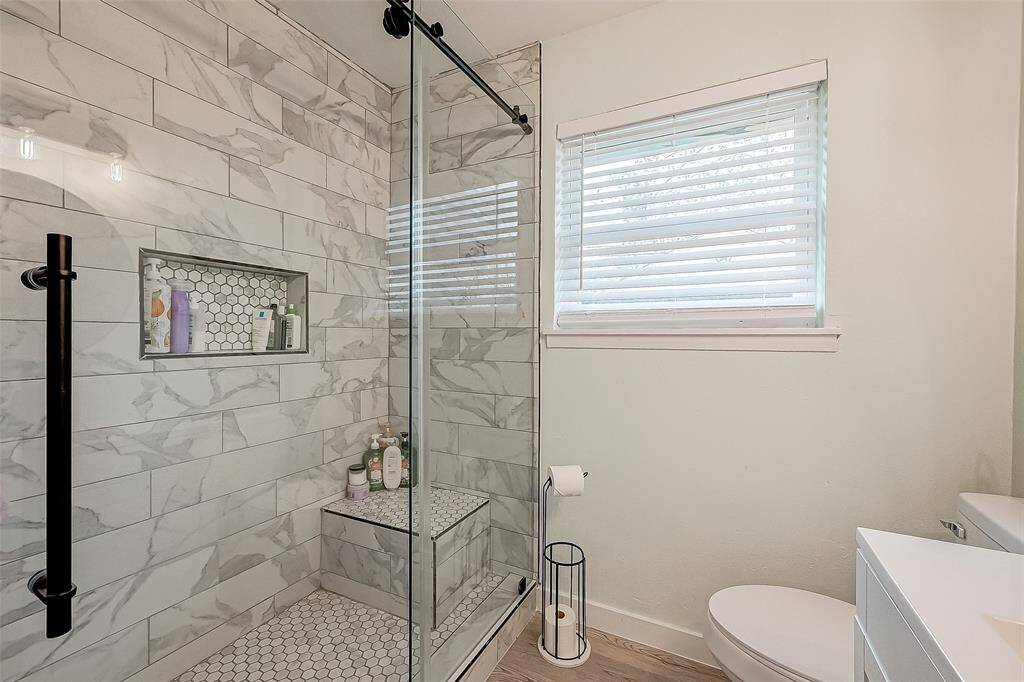 This bathroom features a modern glass-enclosed shower with stylish marble-patterned tiles and a built-in shelf. It has a large window with blinds for natural light and privacy, complemented by a sleek white vanity and toilet.