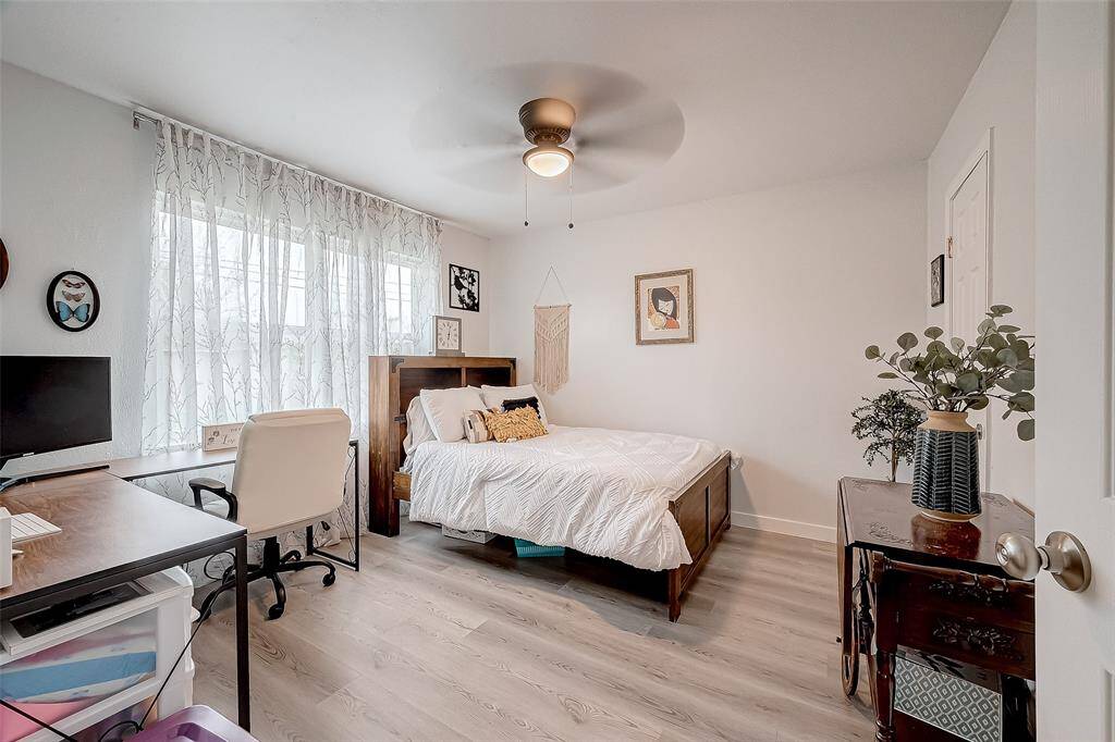 This bright bedroom features a large window with sheer curtains, allowing natural light to fill the space. There's a cozy bed, a ceiling fan, a wooden desk with an office chair, and tasteful decor, creating a welcoming and functional environment.