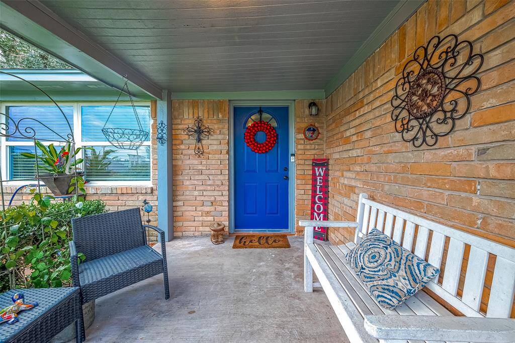 Charming front porch with a vibrant blue door and welcoming decor. Features a cozy seating area with a white bench and wicker chair, surrounded by decorative plants and wall art. Perfect for relaxing and enjoying the neighborhood view.