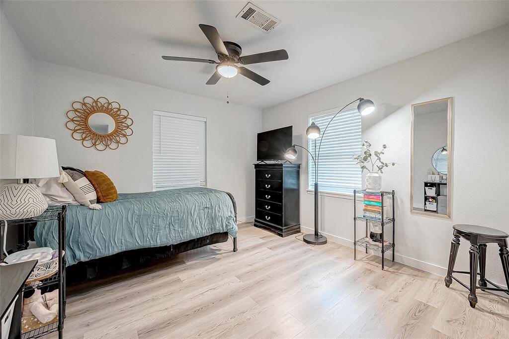 This bedroom features modern decor with light wood flooring and neutral walls. It includes a single bed with cozy bedding, a ceiling fan, and ample natural light from two windows.