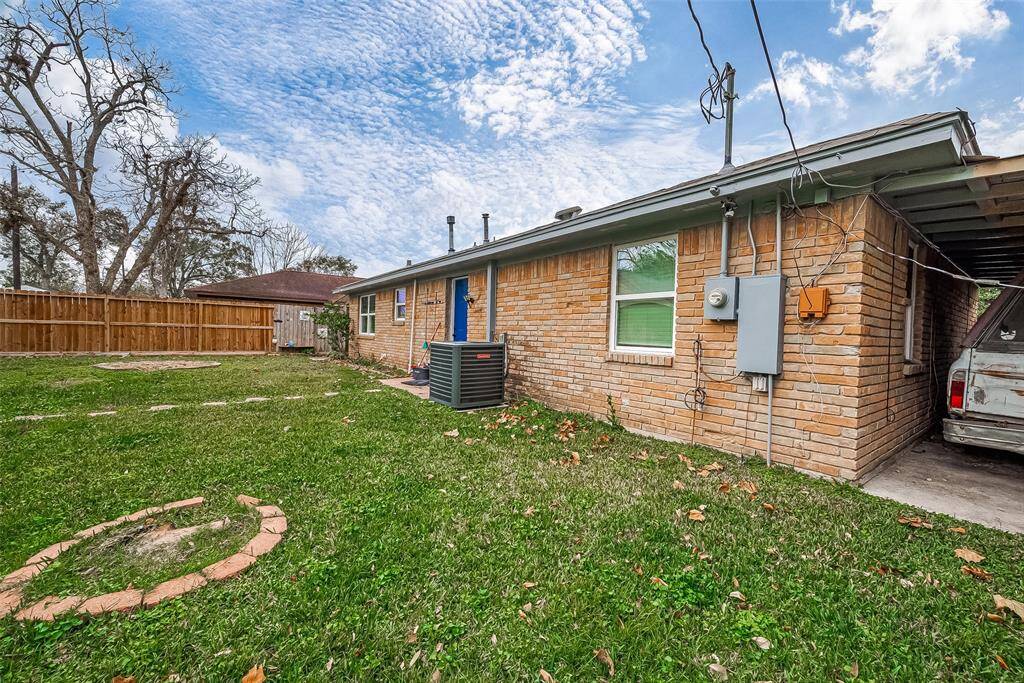 This photo shows the exterior of a single-story brick home with a grassy backyard. There's a wooden fence, some trees, and a carport on the side.