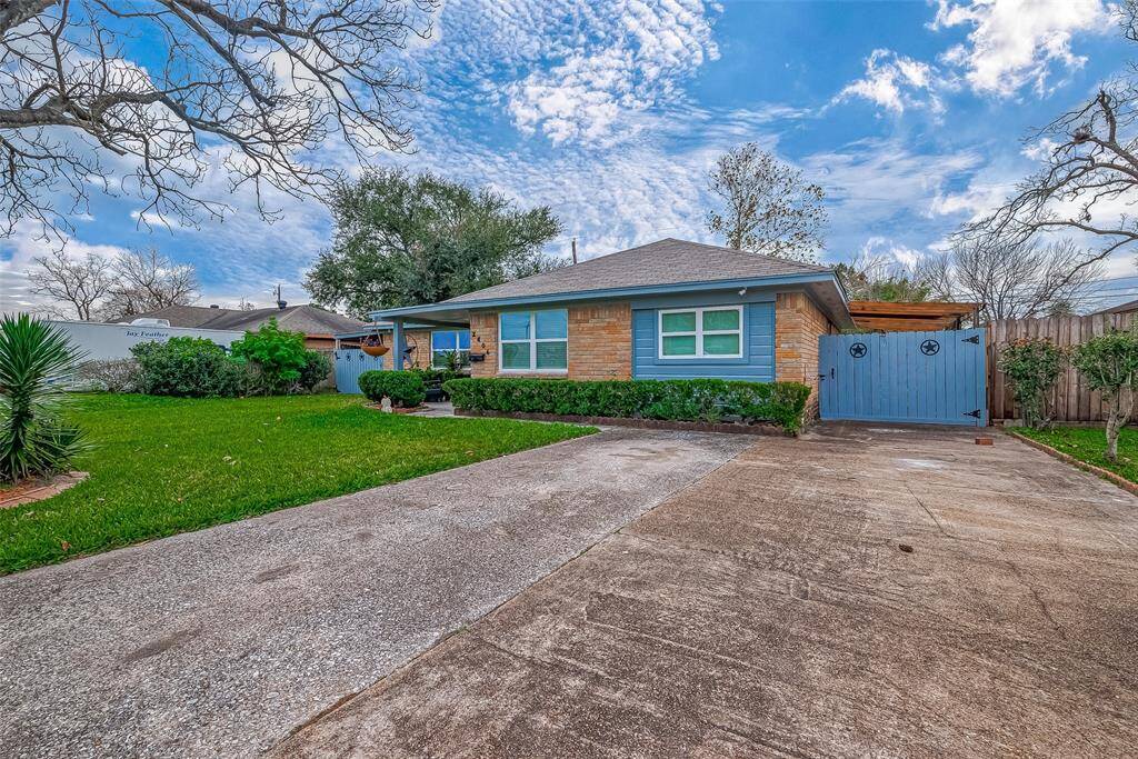 This charming single-story home features a well-maintained front yard with lush greenery and a spacious driveway. The exterior boasts a combination of brick and blue siding, complemented by a wooden gate. The setting is serene, with mature trees and a clear blue sky above.