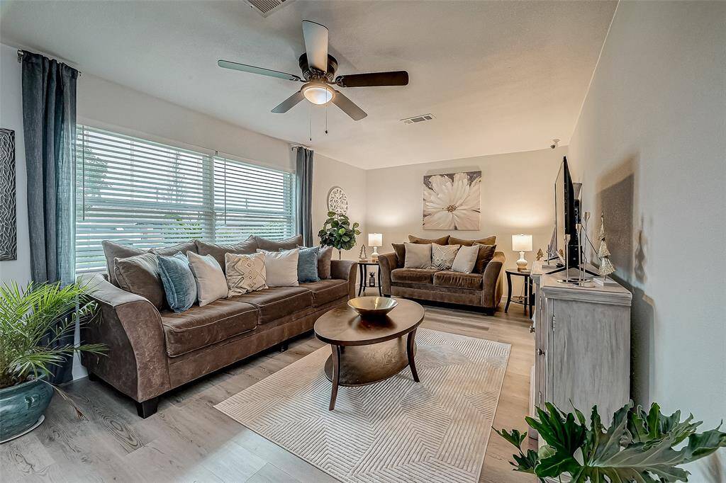 This inviting living room features a cozy setup with large windows with blinds provide ample natural light, complemented by a ceiling fan for comfort. The decor includes a mix of greenery and tasteful artwork, creating a warm and welcoming atmosphere.