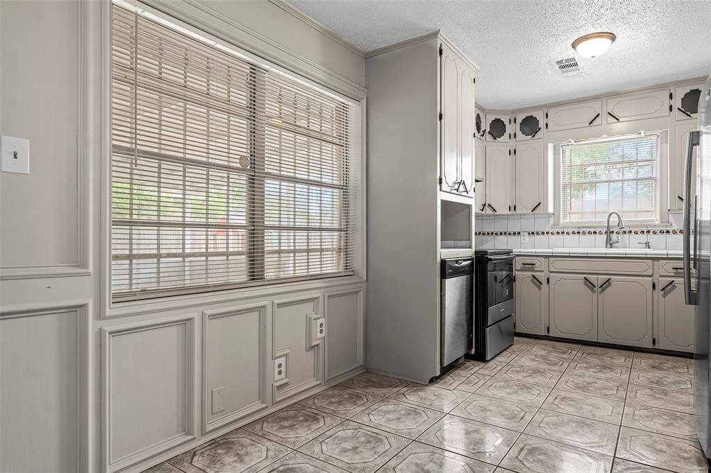 This kitchen features vintage-style cabinetry with decorative details, large windows with blinds for ample natural light, and tiled flooring. It includes modern stainless steel appliances and a cozy, functional layout.