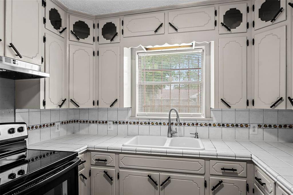 This kitchen features vintage-style cabinetry with decorative handles, a large window above the sink for natural light, tiled countertops, and a classic backsplash with floral accents.