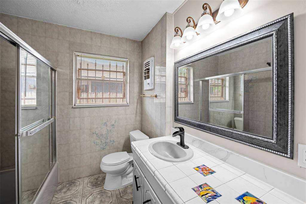 This bathroom features a spacious layout with a glass-enclosed shower, large mirror with decorative frame, and ample natural light from the window. The vintage-style tiles add character, complemented by a classic light fixture above the vanity.