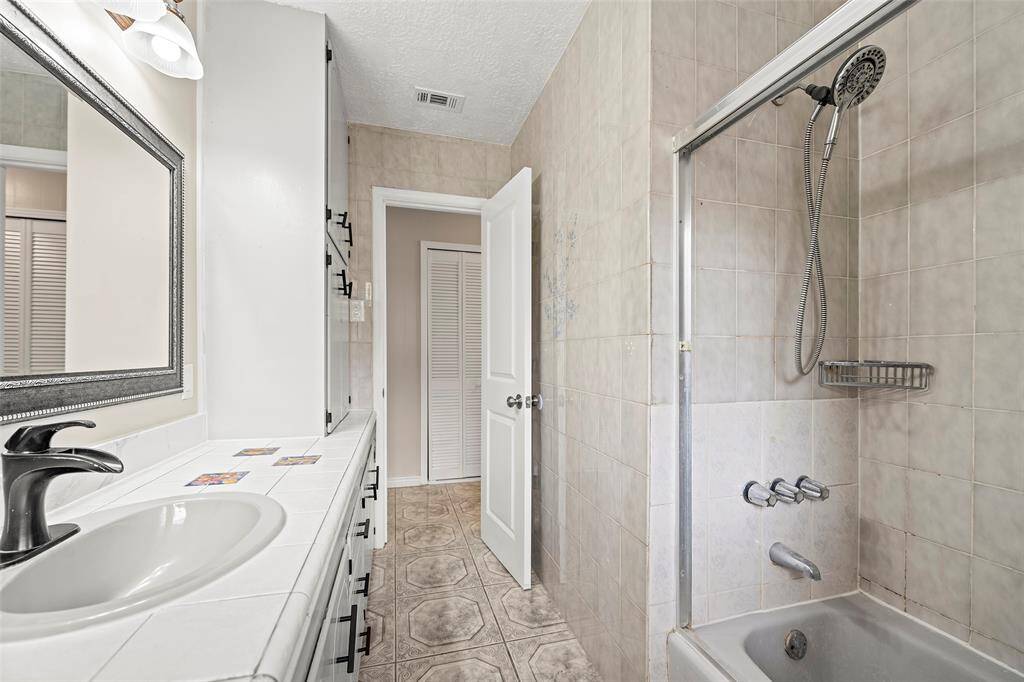 This bathroom features a tiled shower-tub combination, a single-sink vanity with ample counter space, and a large mirror. The neutral color palette and built-in storage create a clean and functional space.