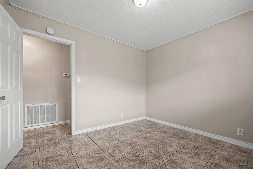 Third bedroom with neutral-painted walls and tiled flooring. It features a single ceiling light, a white door, and a vent.