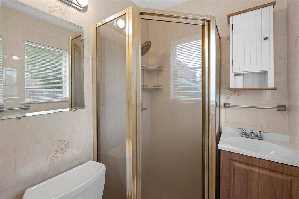 This primary bathroom features a compact design with a glass-enclosed shower, a small vanity with a sink, and a wall-mounted cabinet for storage. The space is brightened by natural light through a window with blinds, and the walls are tiled with a subtle floral pattern.