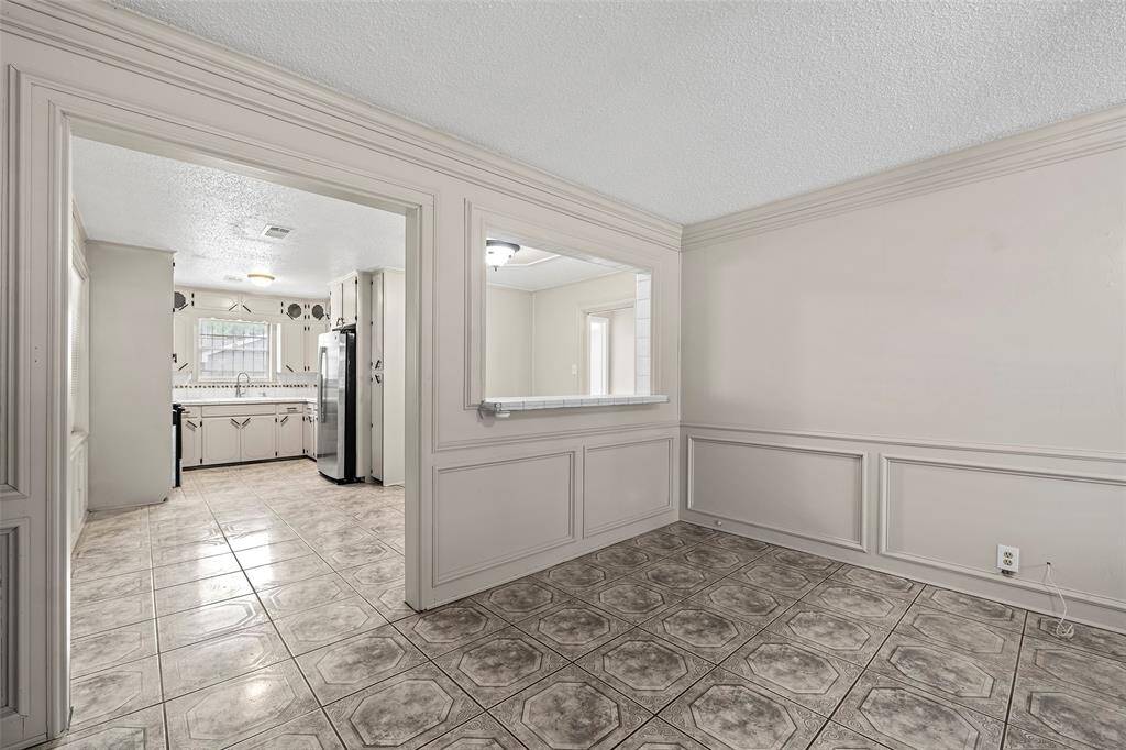 This photo shows a bright room with tiled flooring and neutral walls, featuring a pass-through window to a spacious kitchen. The kitchen includes a stainless steel refrigerator, ample cabinetry, and a large window for natural light.