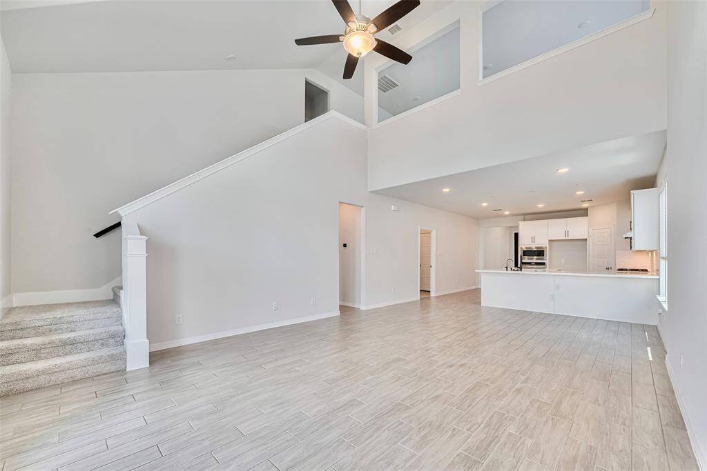 Modern open-concept family room features a high ceiling with a ceiling fan, large windows for natural light. The room has tiled floors and carpeted stairs leading to a second level, offering a bright and inviting living space.