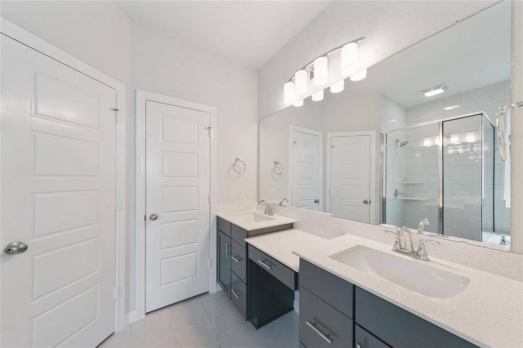 This modern bathroom features dual sinks with sleek fixtures, a large vanity mirror, and a spacious glass-enclosed shower. The room is bright and airy, with contemporary cabinetry and neutral tile flooring.