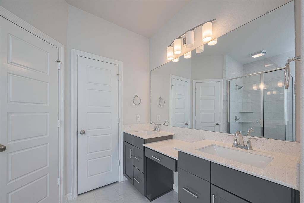 Dual sinks, sleek cabinetry, and a large mirror. It includes a glass-enclosed shower and stylish light fixtures, providing a clean and contemporary feel.
