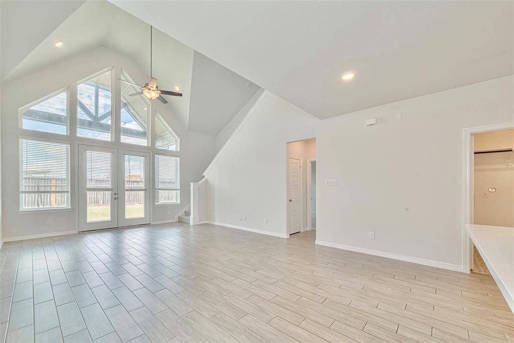 This spacious living room features high vaulted ceilings, large windows for abundant natural light, and sleek tile flooring. A modern ceiling fan complements the bright, open space, leading to a staircase and adjacent rooms. Perfect for a contemporary feel.