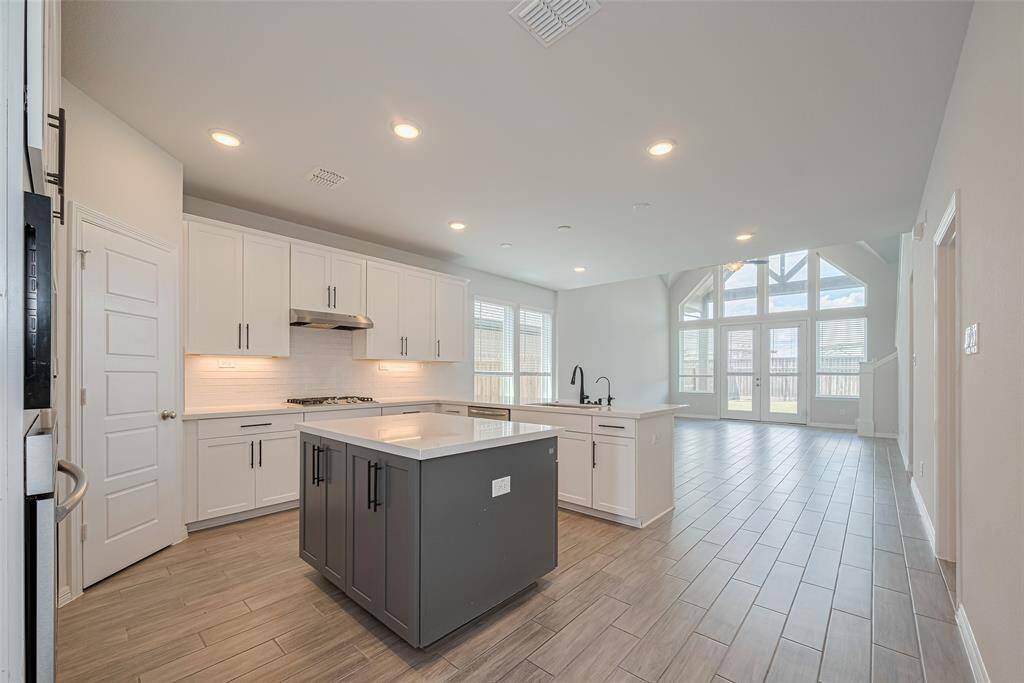 This modern kitchen features sleek white cabinetry, a stainless steel gas stove, and a spacious island with a black faucet. Ample natural light flows in through large windows, creating an inviting and airy atmosphere.