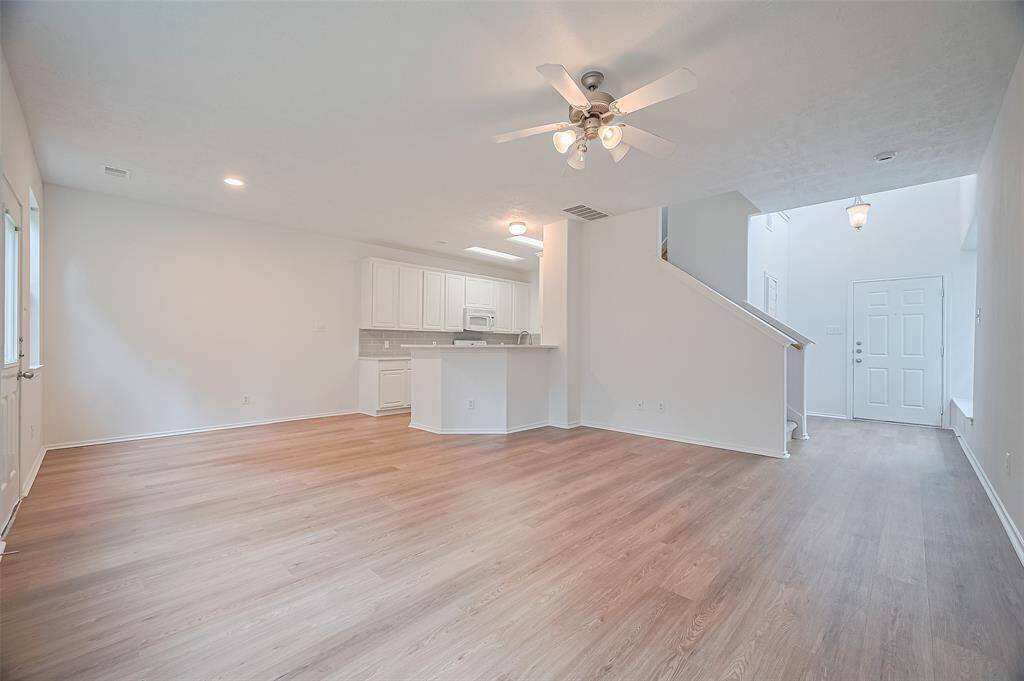 The interior features an open-concept living area with light wood flooring, a ceiling fan, and recessed lighting. The space flows into a modern kitchen with white cabinetry and a breakfast bar. A staircase leads to the upper level, and there's a welcoming entryway with natural light.