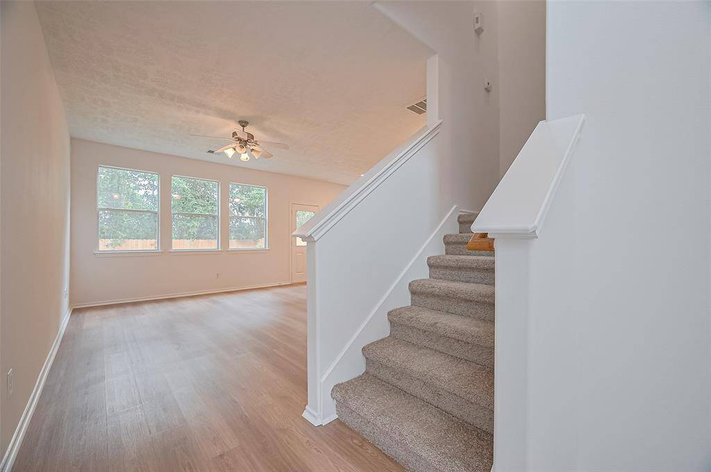 Enjoy a bright living area with large windows, a ceiling fan, and light wood flooring. Carpeted stairs lead to an upper level, offering a spacious and inviting entryway.