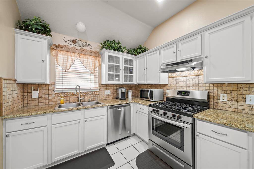 This kitchen features white cabinetry, a gas stove, stainless steel appliances, and granite countertops. The backsplash has a warm, earthy tile design. A window above the sink provides natural light, and plants add a touch of greenery.