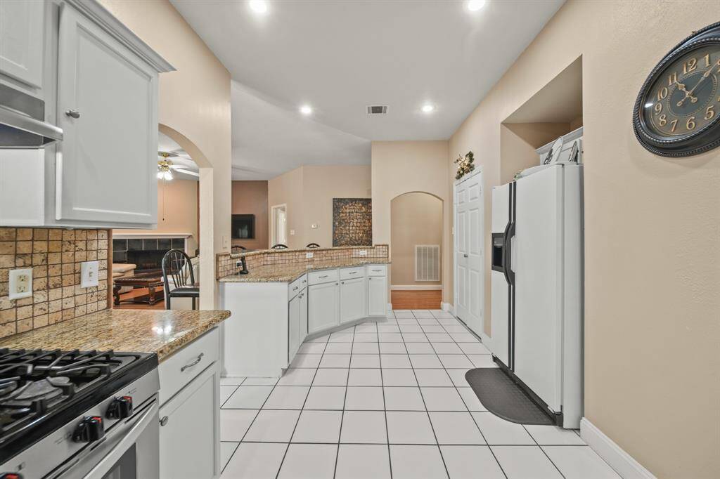 This kitchen features white cabinetry, granite countertops, and a tiled backsplash. It has a spacious layout with tiled flooring, recessed lighting, and a view into a cozy adjacent living area with a fireplace.