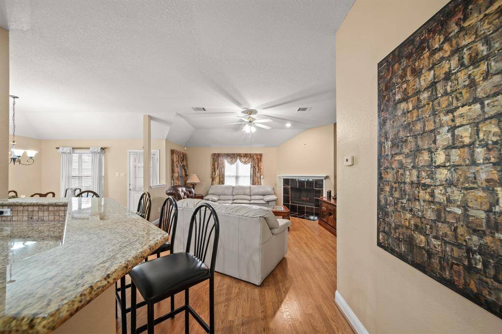 This inviting open-concept living area features hardwood floors, a cozy seating area with a fireplace, and a kitchen with a granite countertop bar. The space is well-lit with natural light and has a ceiling fan for comfort.