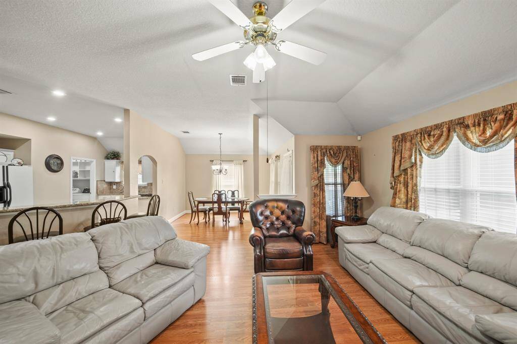 This spacious living area features hardwood floors, a vaulted ceiling with a ceiling fan, and ample natural light. It connects seamlessly to a dining area and kitchen with a breakfast bar, creating an open and inviting space perfect for entertaining.