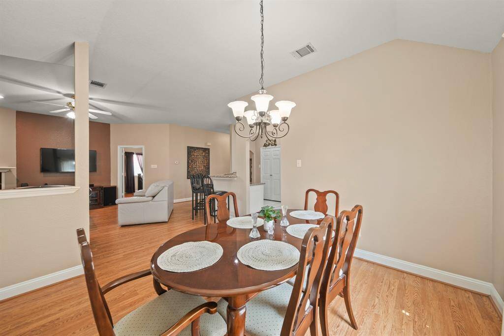 This photo showcases a bright, open dining and living area with hardwood floors, neutral walls, a stylish chandelier, and a view into the living room with a ceiling fan and TV. Ideal for entertaining and family gatherings.