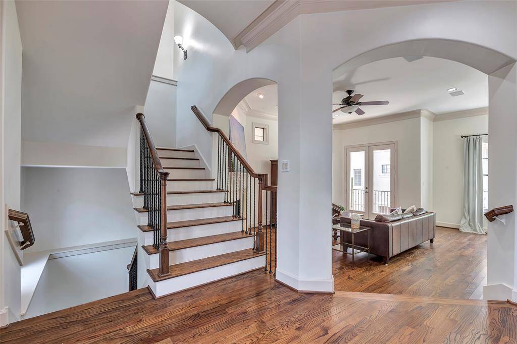 This photo showcases a bright and spacious interior with hardwood floors, a staircase with elegant wrought iron railings, and an open-concept living area. The room features large windows, a ceiling fan, and French doors, providing a modern and inviting atmosphere.