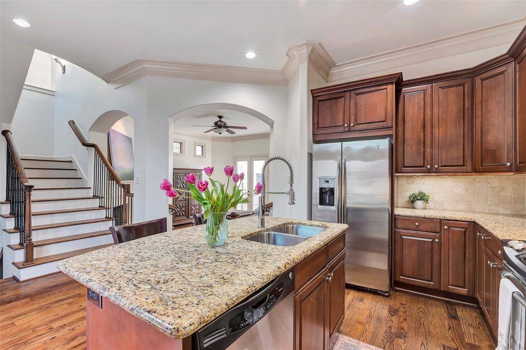 This sophisticated kitchen has rich wooden cabinetry, granite countertops, and stainless-steel appliances. It features a central island with a sink, an adjacent open staircase with dark railings, and an arched entryway leading to a bright living area.