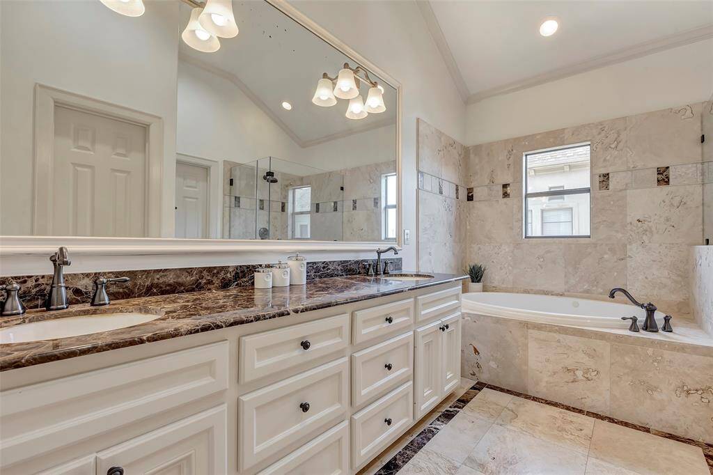 This bathroom is spacious and elegant, featuring a luxurious double vanity with marble countertops, a large mirror, and stylish light fixtures. The room includes a relaxing bathtub set against a tiled wall, with a separate glass-enclosed shower. Natural light filters in through a window, enhancing the serene atmosphere.
