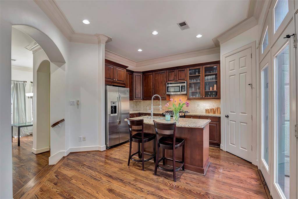 This kitchen features elegant wood cabinetry, granite countertops, and stainless steel appliances. It has a central island with seating for two, hardwood floors, and ample natural light from large windows, creating a warm and inviting space.