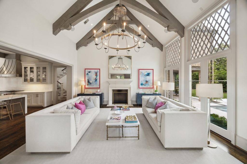 Elegant living room with vaulted, beam-accented ceilings, statement lighting, and floor-to-ceiling windows overlooking the pool. Open to the kitchen and adjacent to the back staircase for seamless flow.